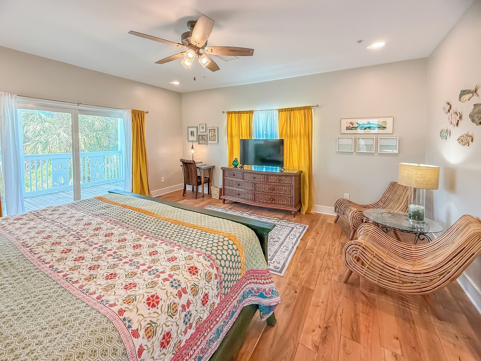 A spacious bedroom with a large bed, a ceiling fan, a TV, and a balcony with yellow curtains.