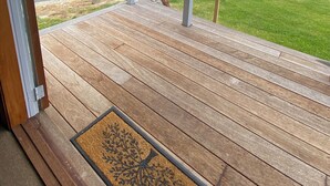 Eine Holzterrasse mit Fußmatte, Blick auf eine grüne Landschaft mit Hügeln und Bäumen, und ein bewölkter Himmel.