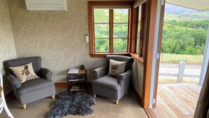 Ein gemütliches Wohnzimmer mit zwei grauen Sesseln, einem kleinen Couchtisch und einem schwarzen Teppich. Ein Fenster bietet Blick auf grüne Hügel.