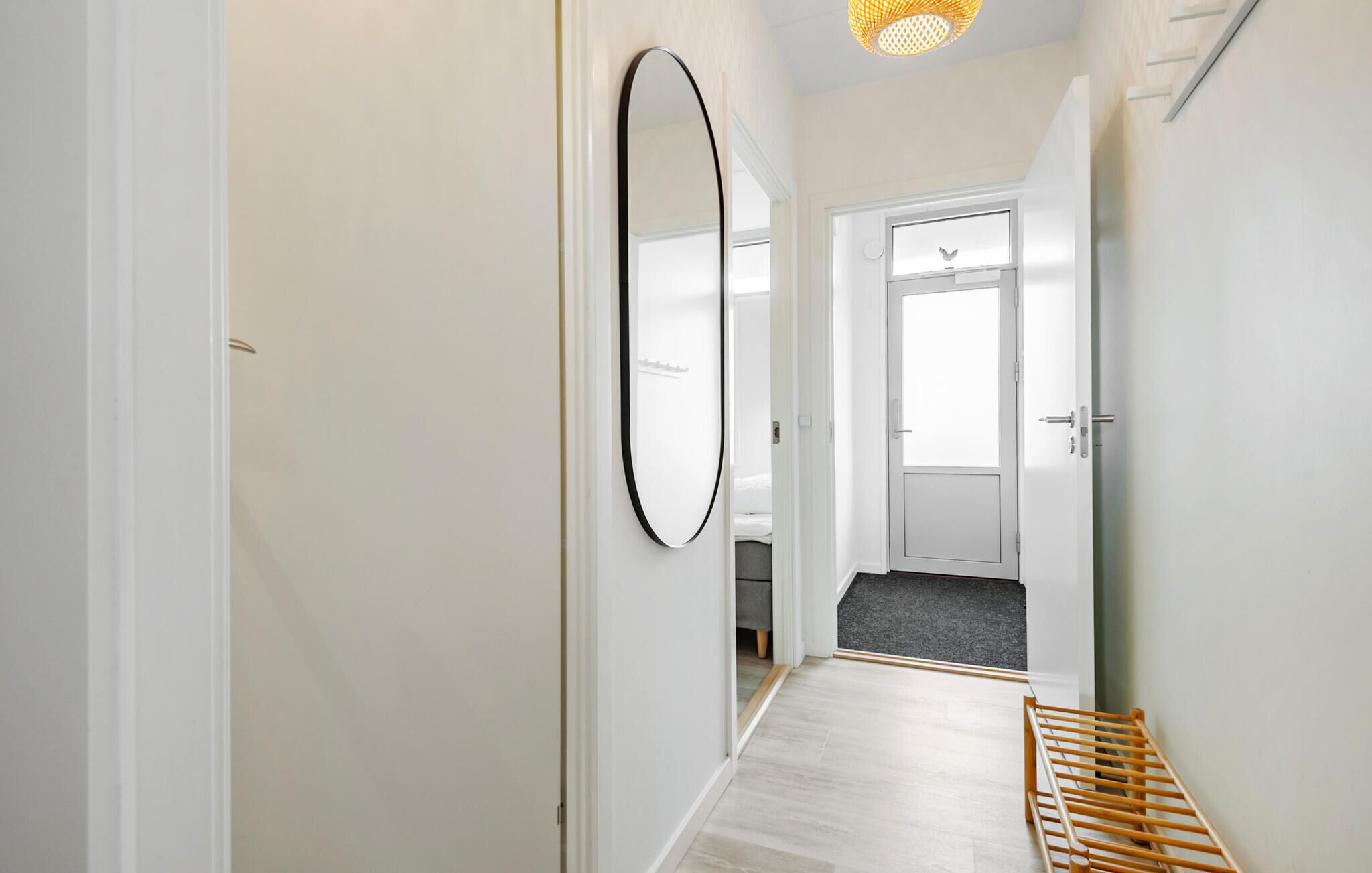 Un couloir avec un banc en bois, un grand miroir et une porte donnant sur une pièce où se trouve un lit.