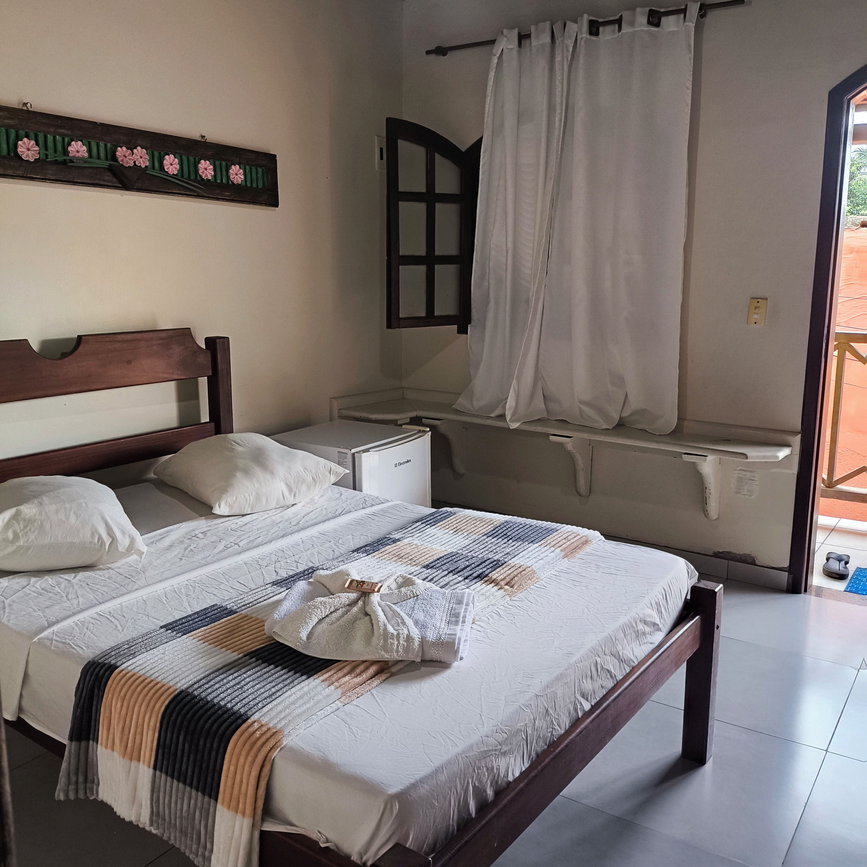 Una habitación de hotel con cama, mesita de noche, ventana con cortinas y estante fijado a la pared.