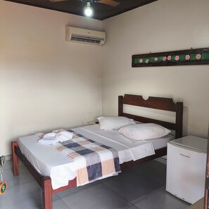 Una cama individual con cabecera de madera, una mesita de noche, un refrigerador blanco pequeño y un equipo de aire acondicionado en la pared.