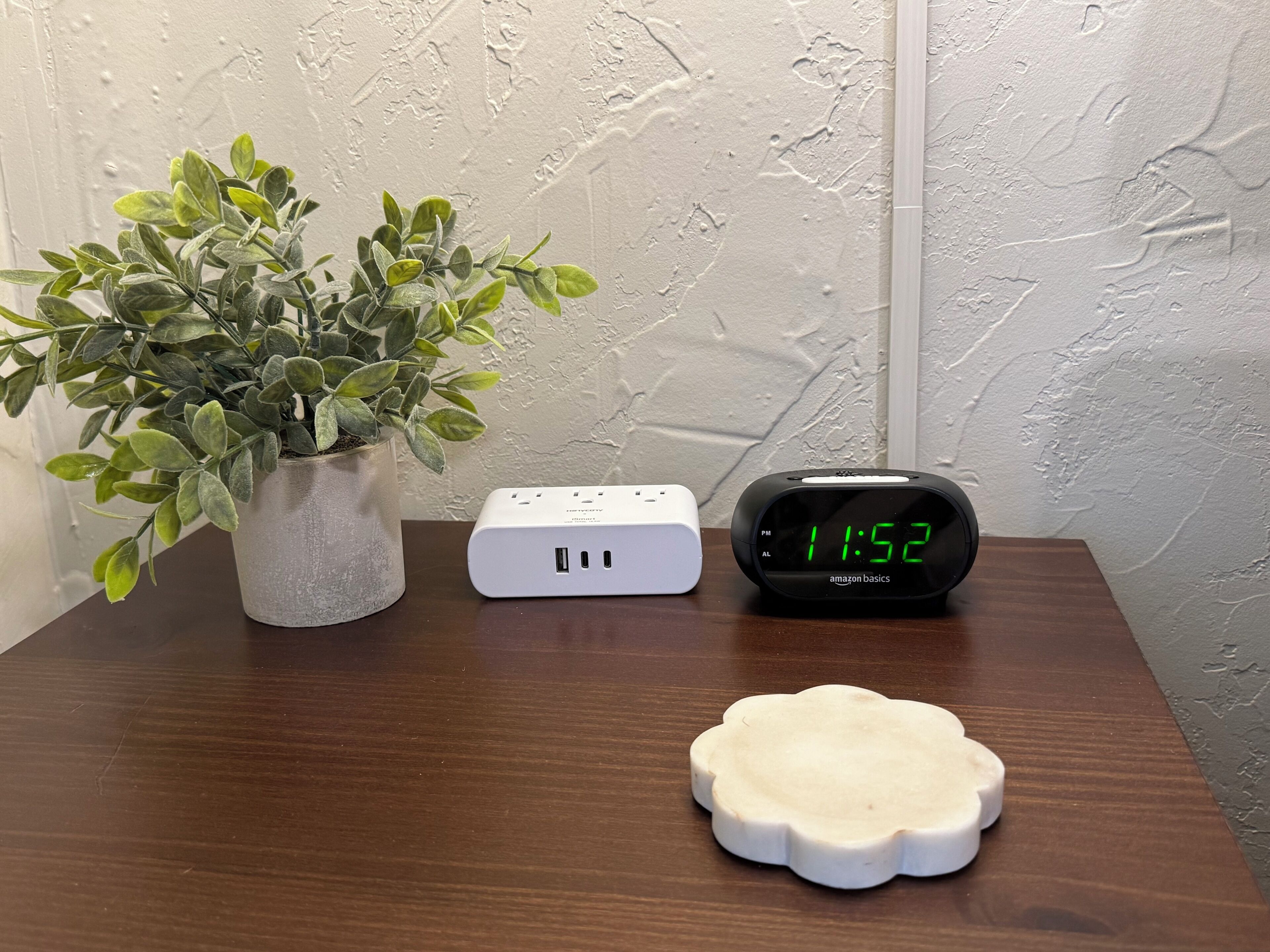 A wooden table with a potted plant, a white power strip, and a black digital clock displaying 11:52.