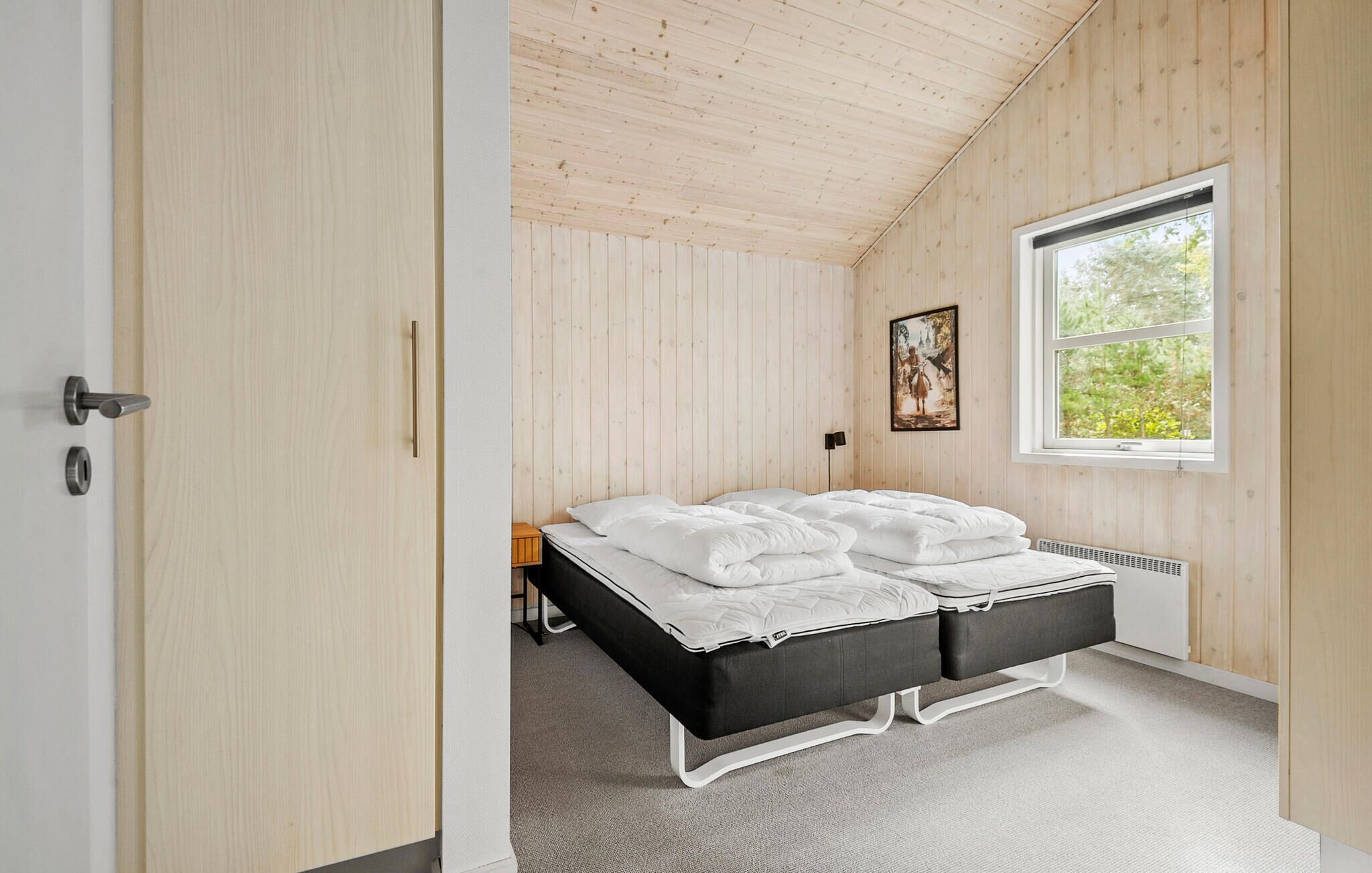 A bedroom with a wooden ceiling, a bed with white bedding, a window with a view of greenery, and a painting on the wall.