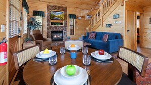 A cozy cabin interior with a dining table set for four, a fireplace, a blue sofa, and a staircase. - Bear's Den |  Hot Tub~Fireplace~BBQ~Sleeping Loft (Overgaard)