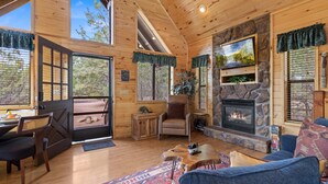 A cozy cabin interior with a stone fireplace, wooden walls, and a large window overlooking a forest. - Bear's Den |  Hot Tub~Fireplace~BBQ~Sleeping Loft (Overgaard)