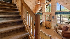 A wooden cabin interior with a staircase, a ceiling fan, a dining area, and a view of the outdoors. - Bear's Den |  Hot Tub~Fireplace~BBQ~Sleeping Loft (Overgaard)