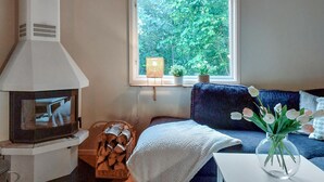 Een gezellige woonkamer met een open haard, een raam met uitzicht op groen, een blauwe bank en een vaas met bloemen op een tafel.