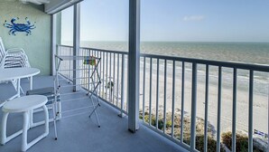 A balcony with a view of the ocean, a white table, a white chair, and a metal drying rack. - Sandcastle I 907—Top-Floor Gulf-Front Condo w/ Panoramic Views, Pool, & Modern Upgrades (Indian Shores)