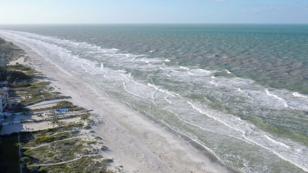 A beach with a sandy shore, a few buildings, and a flag. - Sandcastle I 907—Top-Floor Gulf-Front Condo w/ Panoramic Views, Pool, & Modern Upgrades (Indian Shores)