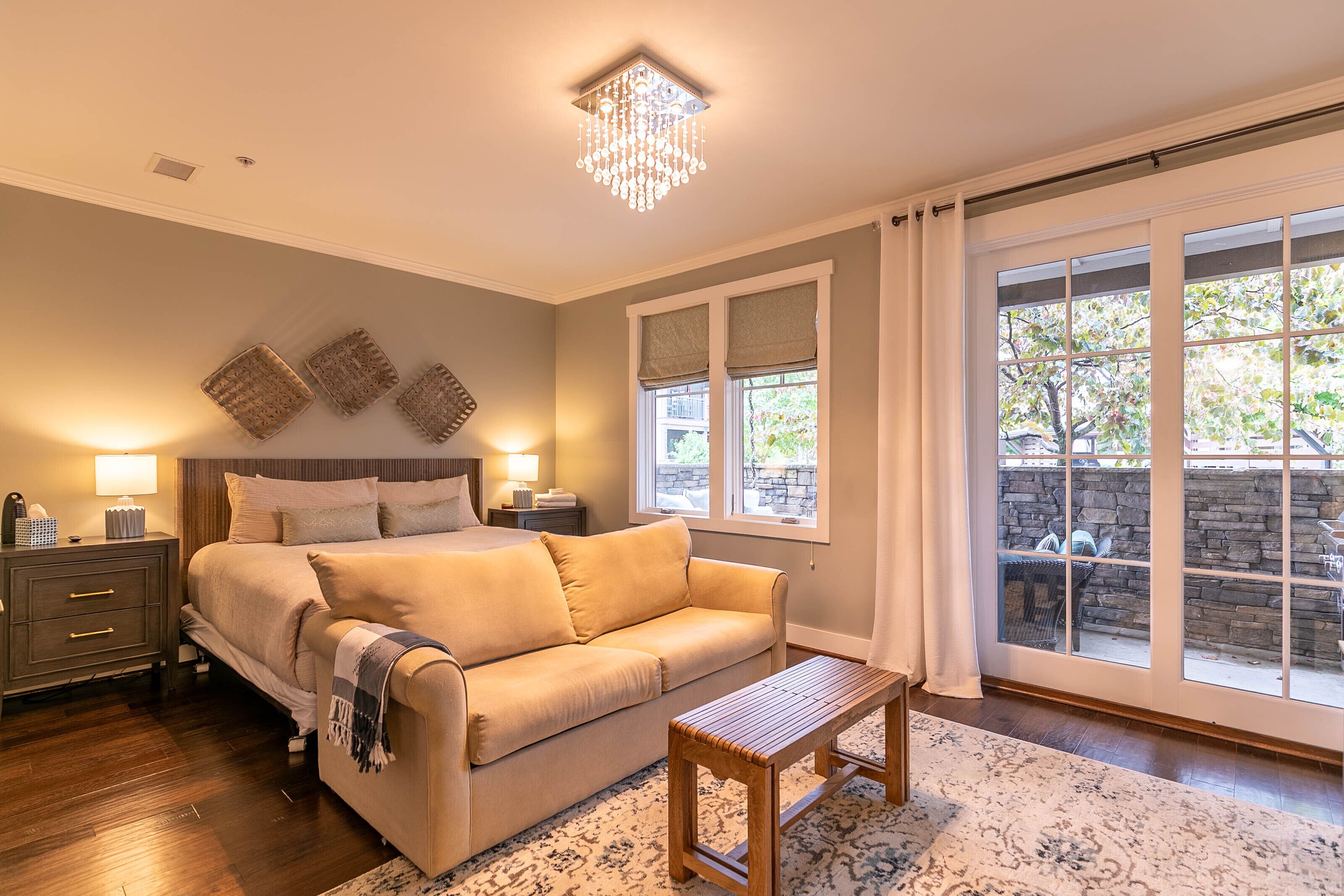 A bedroom with a bed, a sofa, a nightstand, a lamp, and a large window with a view of a stone wall and greenery.