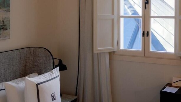 Una habitación de hotel con cama, ventana con cortinas, un cuadro en la pared y una mesita de noche con lámpara.