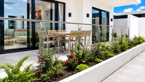 Un patio exterior moderno con puerta de vidrio que da a un área de estar, una mesa y sillas de madera, y diversas plantas.