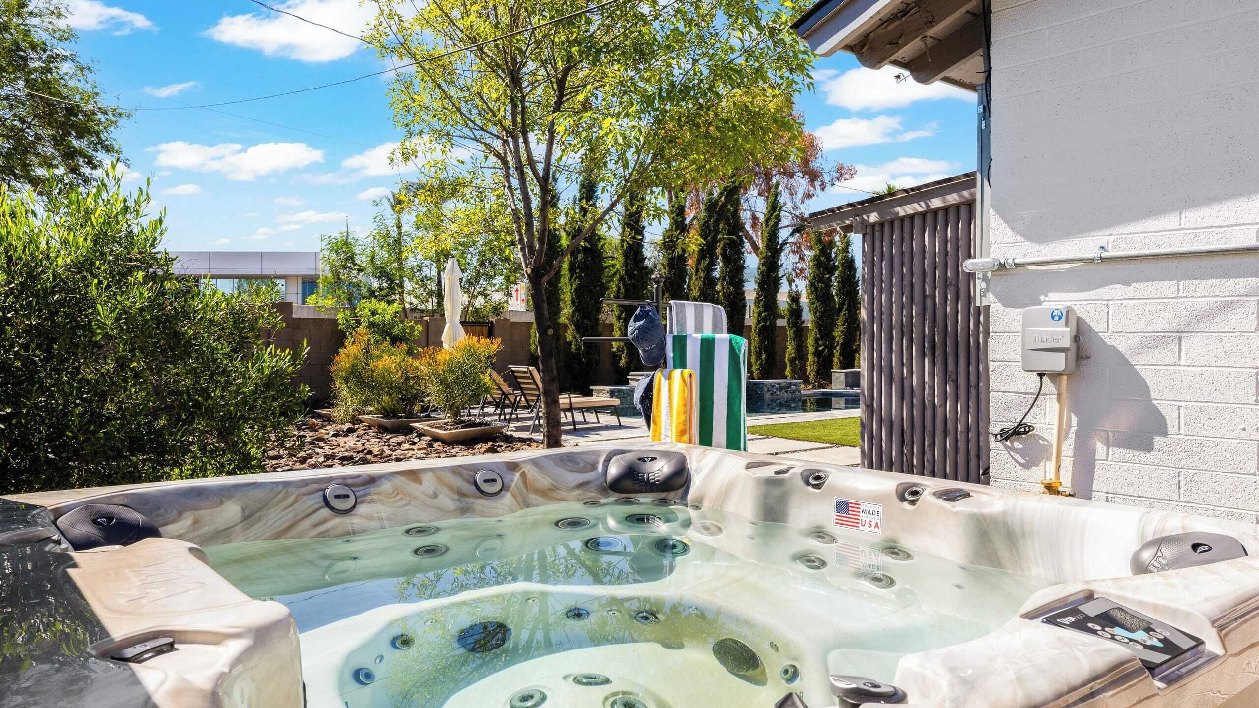 Un bain à remous doté de plusieurs jets, entouré d’un jardin aménagé avec des arbres et des arbustes, et une clôture en bois en arrière-plan.