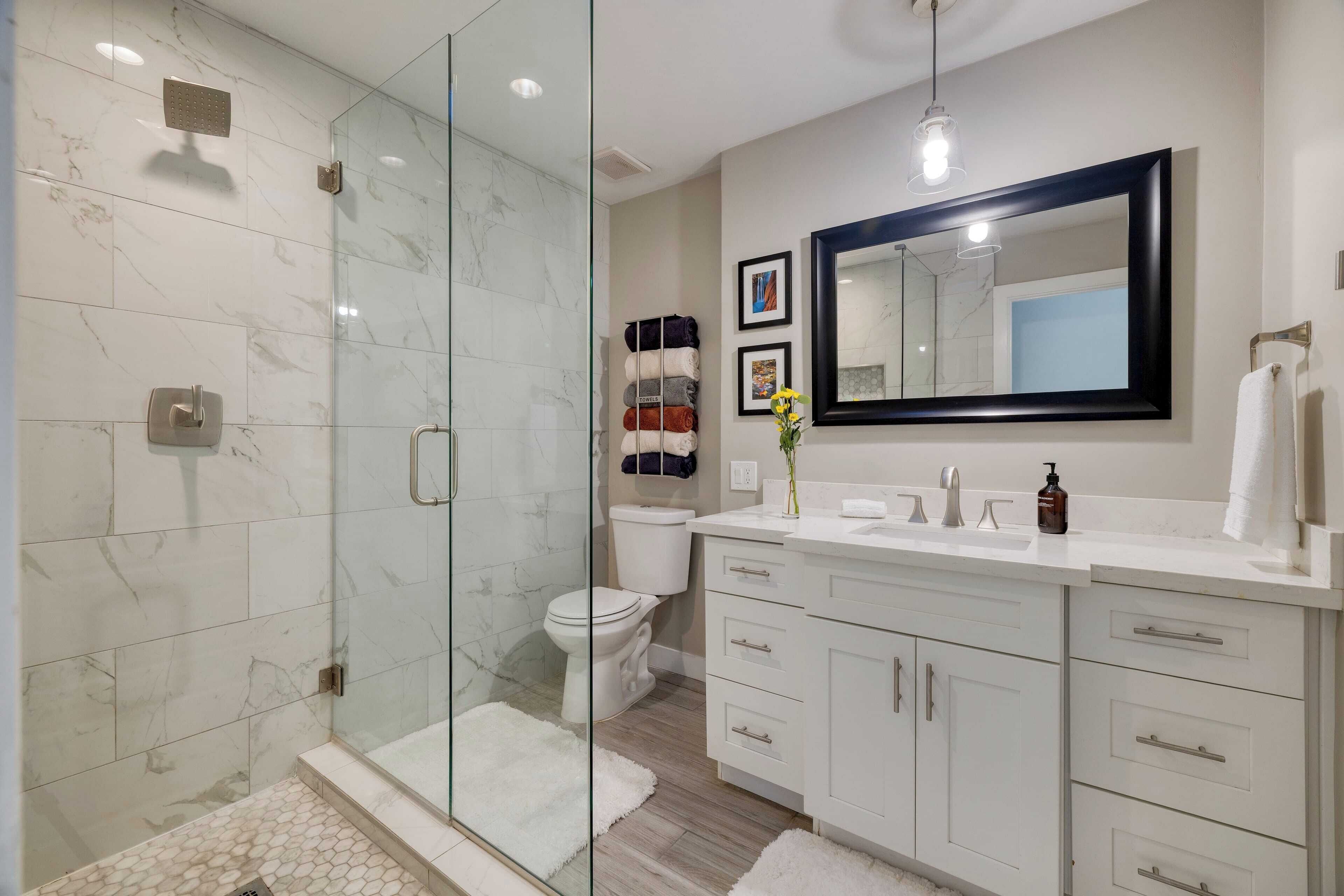 Ein modernes Badezimmer mit einer glasumrandeten Dusche, weißen Marmorfliesen, einem weißen Waschbecken mit großem Spiegel und einer Toilette mit Handtuchhalter.