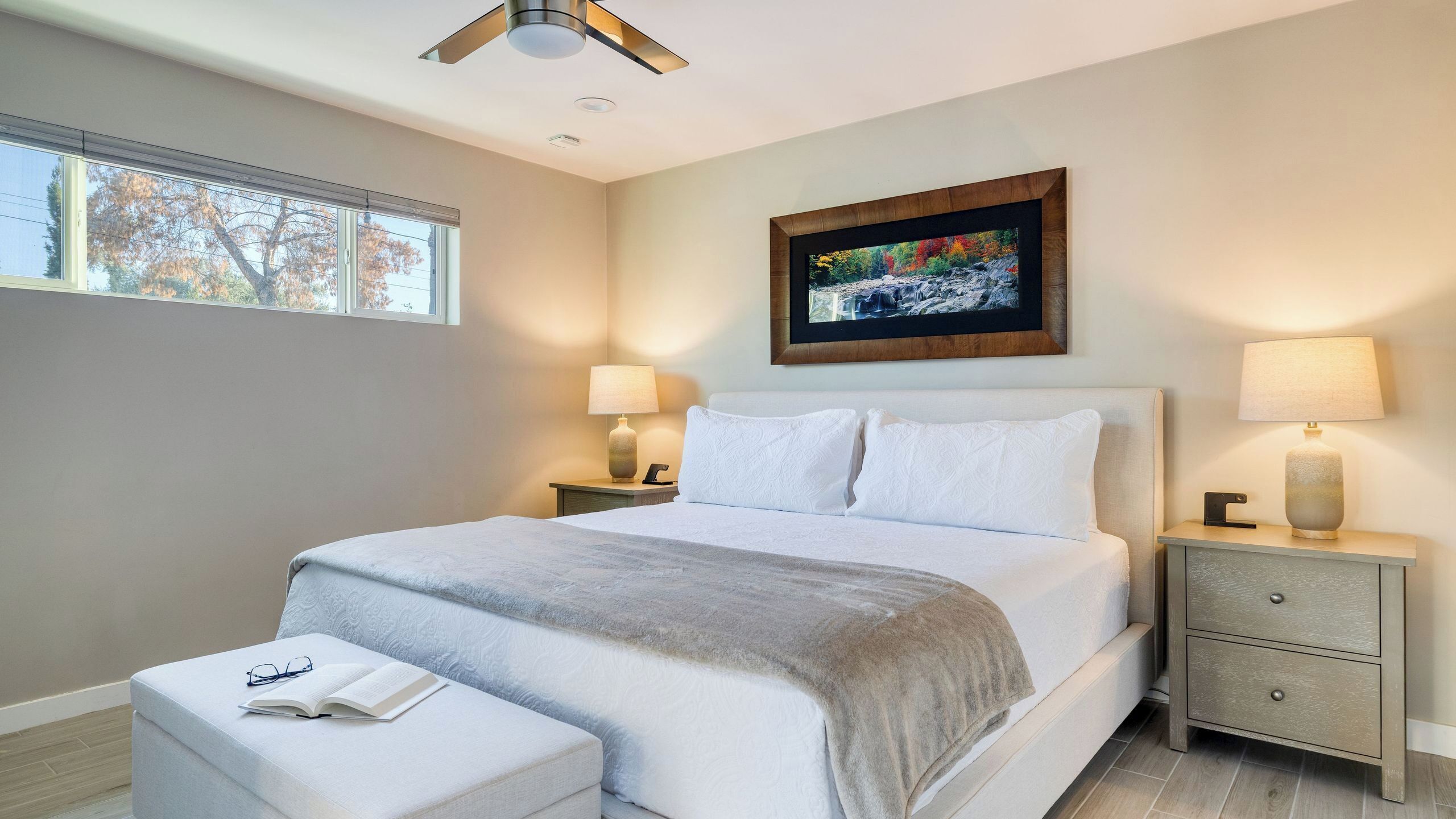 A modern bedroom with a large bed, bedside lamps, a nightstand, a reading chair, and a ceiling fan.