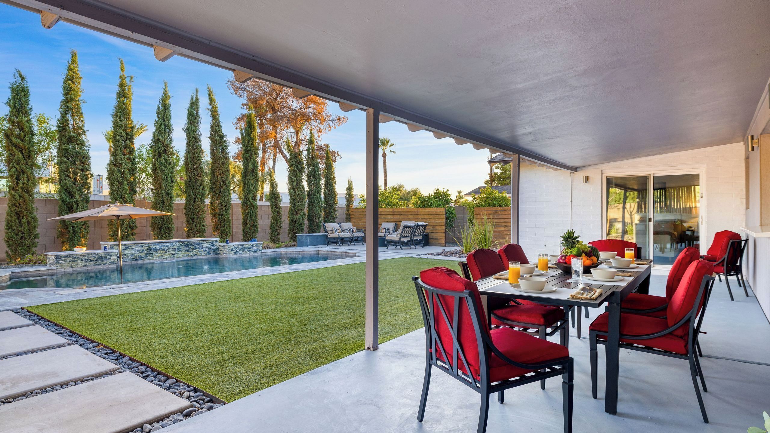 A covered patio with a pool, red chairs, and a dining table.