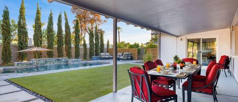 A covered patio with a pool, red chairs, and a dining table.