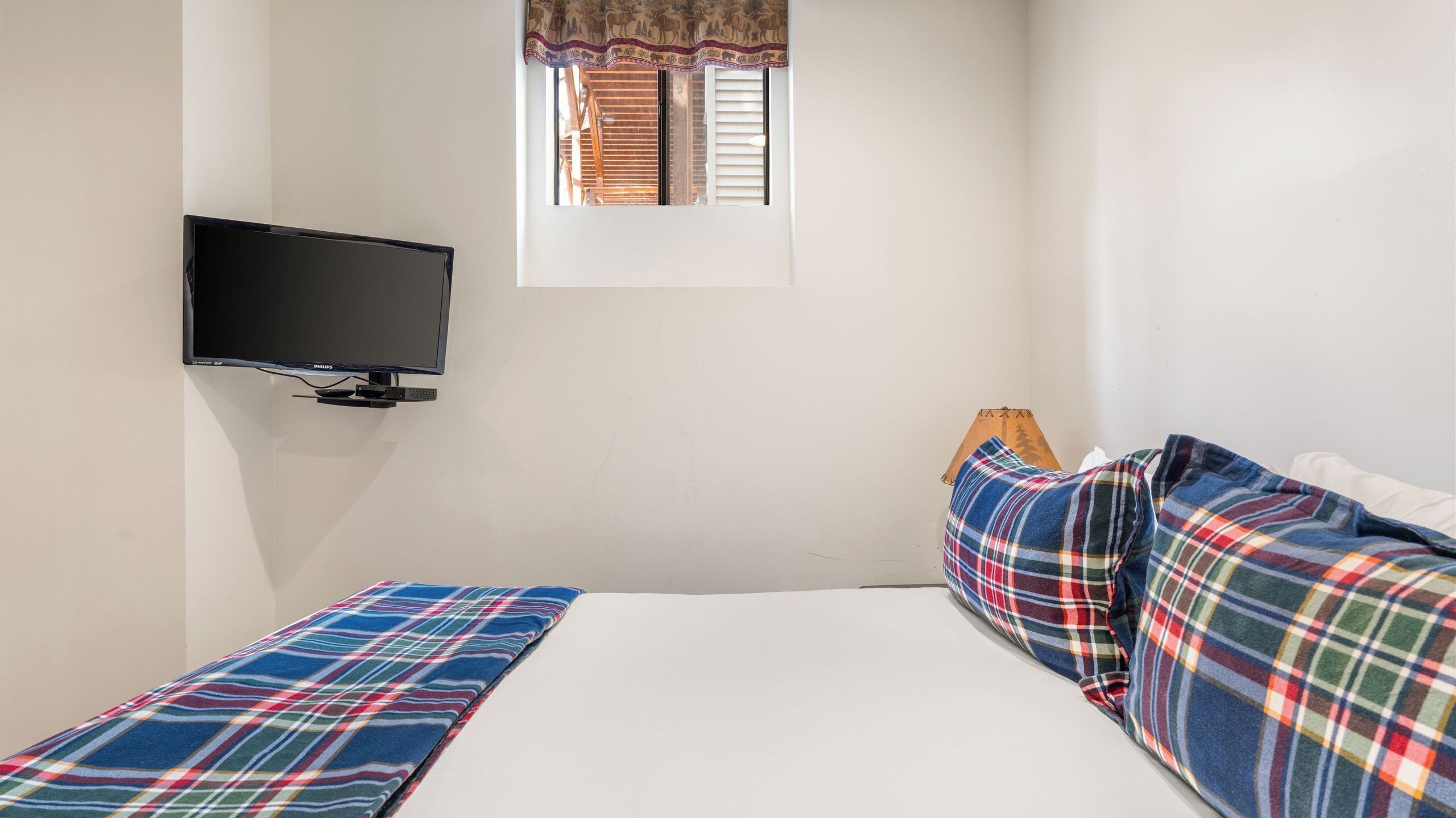A small, single-bed room with a television mounted on the wall, a window with patterned curtains, and a plaid blanket on the bed.