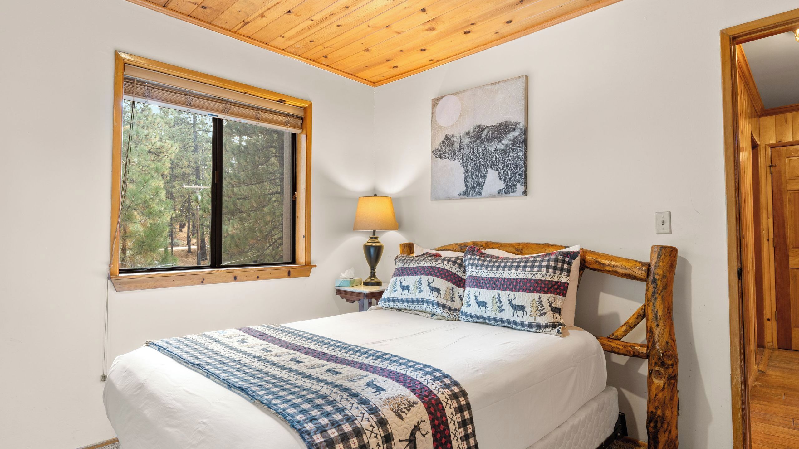 A bedroom with a wooden ceiling, a large window with blinds, a bed with patterned bedding, a bedside lamp, and a wall-mounted painting.