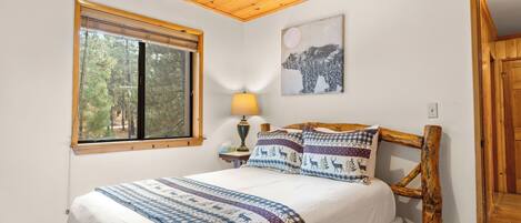 A bedroom with a wooden ceiling, a large window with blinds, a bed with patterned bedding, a bedside lamp, and a wall-mounted painting.