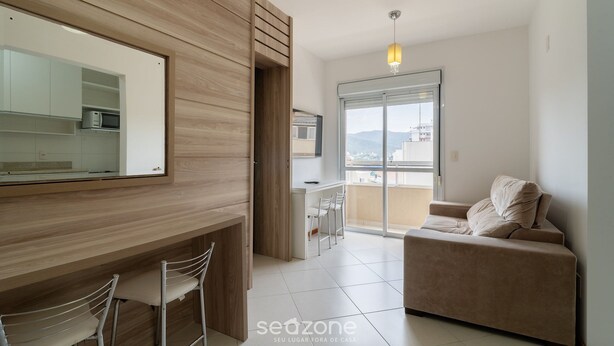 A modern living room with a sofa, a dining area with chairs, a kitchen with a microwave, and a large window offering a view. - 1-bedroom Apartment 850 m From Masc - Cfe0503 (Florianópolis)