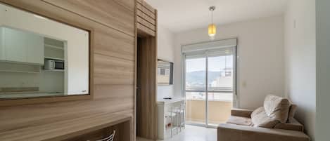 A modern living room with a sofa, a dining area with chairs, a kitchen with a microwave, and a large window offering a view.