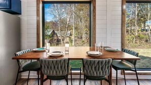 Una mesa de comedor con cuatro sillas, techo de madera y ventanas amplias con vista a árboles y una casa.