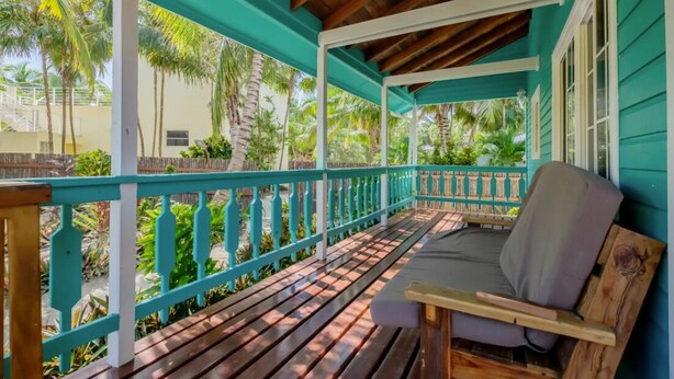 Een overdekte veranda met een houten bank en een beklede zitplaats, omringd door een turquoise hekwerk en palmbomen.