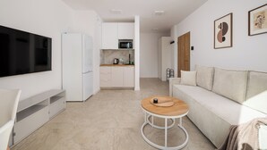 Un espai de vida modern amb televisió de pantalla plana, un sofà blanc, una taula de centre de fusta, una zona de cuina amb armaris blancs i un microones.