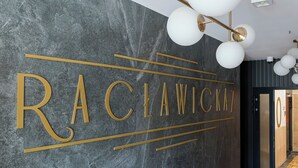 Un interior modern amb una paret de marbre que mostra el nom "RACLAWICH7" amb lletres daurades, una làmpada d'estil contemporani i una porta de fusta amb un logotip circular.