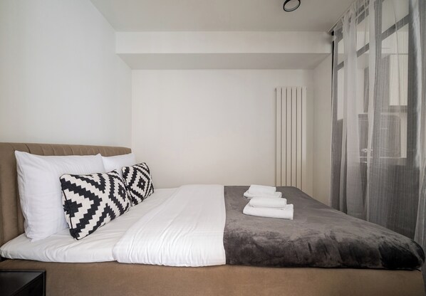 Una cama bien tendida con ropa de cama blanca y gris, una almohada estampada y una toalla doblada al lado.