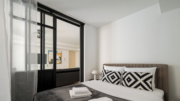 Una habitación de hotel moderna con una cama grande, dos almohadas, una mesita de noche y una ventana con cortinas translúcidas.