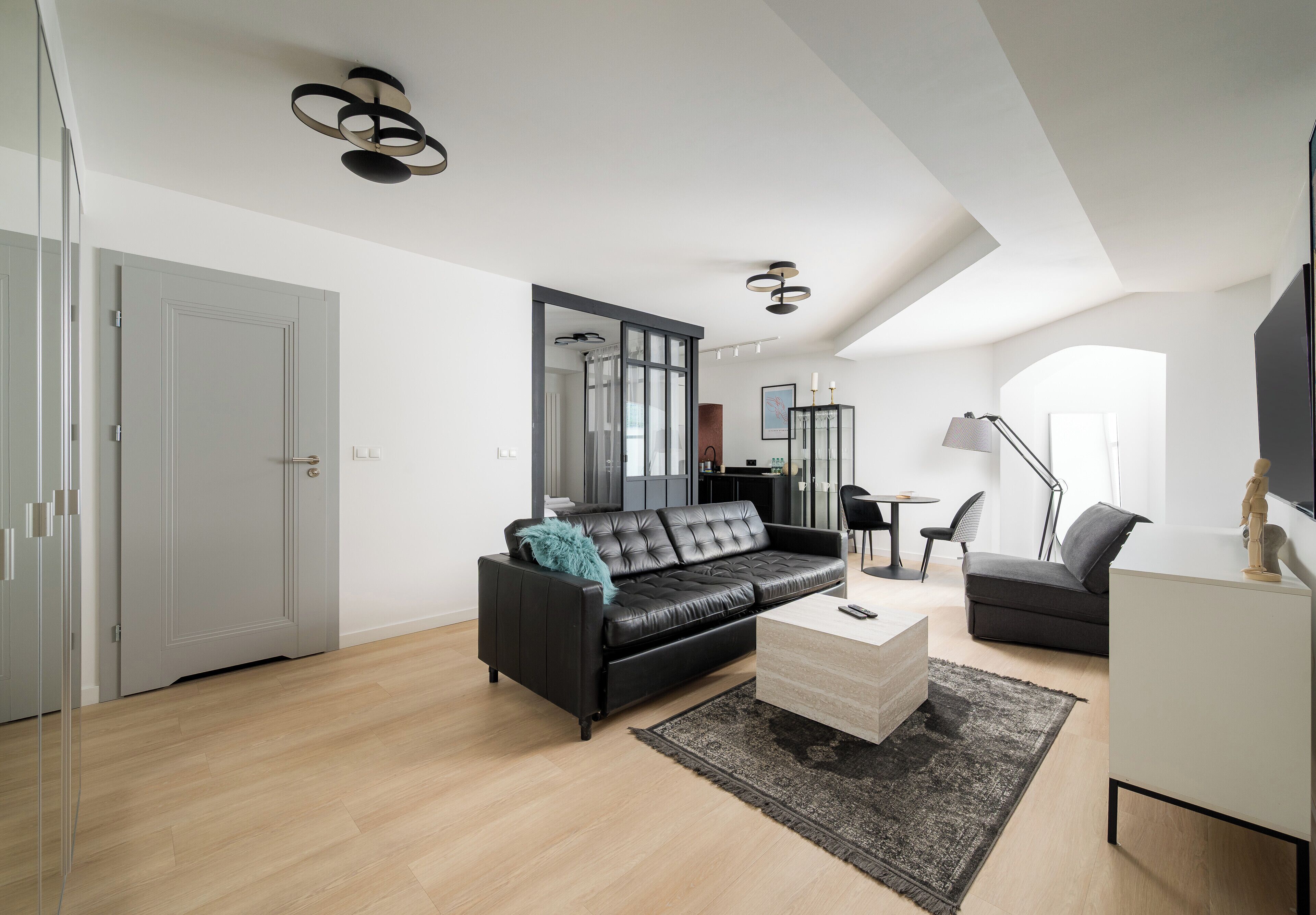 Una sala moderna con un sofá de cuero negro, una mesa de centro blanca y un sillón gris. Hay un espejo grande en la pared, una lámpara de pie negra y un mueble blanco.