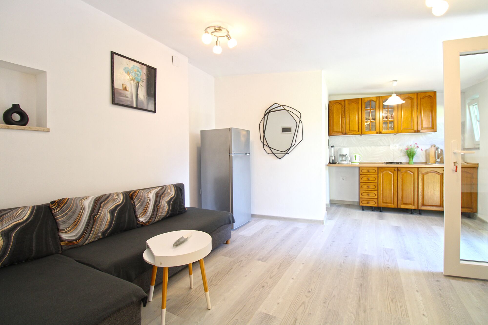 Een moderne woonkamer met een grijze bank, een kleine witte tafel en een grijze koelkast. De keuken heeft houten kastjes en een hangende lamp.