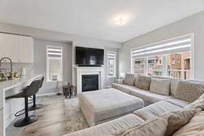 A modern living room with a fireplace, a sofa, a dining area with bar stools, and a TV. - The Devineridge Executive House (Ajax)
