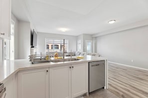 A modern kitchen with white cabinetry, a built-in dishwasher, and a large window providing natural light. - The Devineridge Executive House (Ajax)