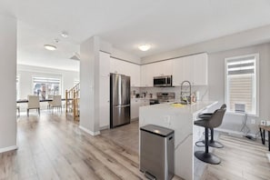 A modern kitchen with stainless steel appliances, a central island with bar stools, and a dining area with a view. - The Devineridge Executive House (Ajax)