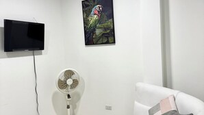 A modern living room with a white sofa, a glass coffee table, a mounted TV, a standing fan, and a framed artwork on the wall. - Cozy apartment (Leticia)