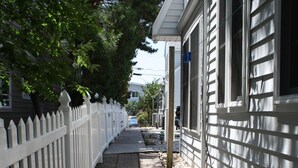 Un trottoir étroit bordé d’une clôture blanche en bois et d’un bâtiment à la façade blanche.