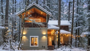 A two-story cabin with a front porch, surrounded by a snowy forest.