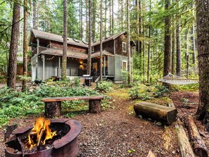Una cabaña en un bosque con un brasero, mobiliario exterior y una hamaca.