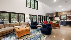 A spacious living area with a large sofa, a wooden coffee table, and a dining table with chairs. There is a kitchen area with a colorful backsplash and a large window overlooking a forest.