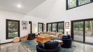 A spacious living room with a large sectional sofa, a wooden coffee table, and a blue armchair. There are large windows offering a view of the forest.