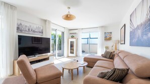 A modern living room with a flat-screen TV, beige sofas, a coffee table, and a large window with a view of greenery.