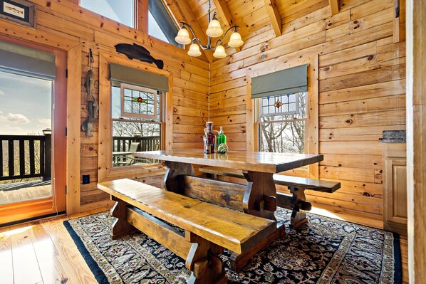 A wooden cabin interior with a large wooden table and benches, large windows with stained glass, and a view of the outside.