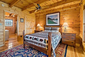 A wooden cabin bedroom with a bed, two nightstands, a ceiling fan, and a large window.