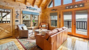A wooden cabin interior with a large sofa, two armchairs, a coffee table, and a view of the outdoors through large windows. - Nice Views, Log Cabin, Gated Community - Mountain Vista by Carolina Properties (Bostic)