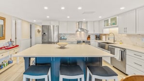 Una cocina moderna con armarios blancos, una isla grande con taburetes, electrodomésticos de acero inoxidable e iluminación empotrada en el techo.