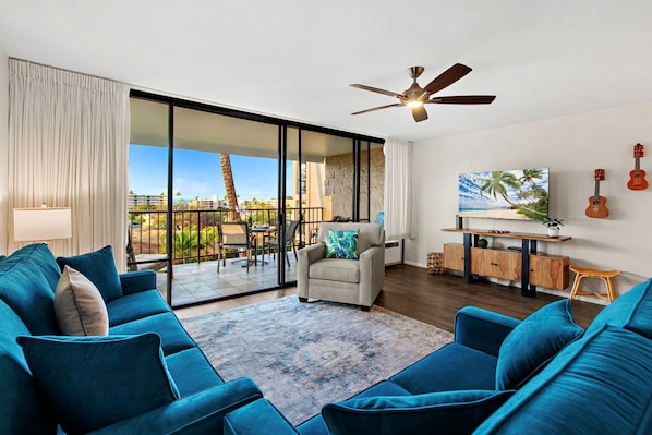 A spacious living room with a large sliding glass door leading to a balcony, a ceiling fan, a TV mounted on a wooden cabinet, and a view of palm trees and buildings. - Recently updated spacious 1bd in Maui (Kihei)
